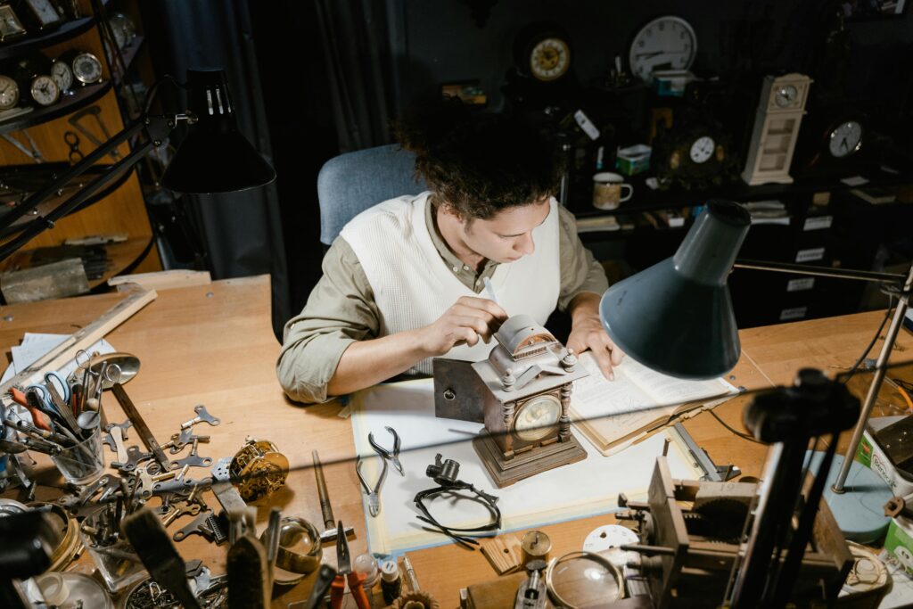 A watchmaker carefully repairs a clock on a cluttered workbench surrounded by tools.
